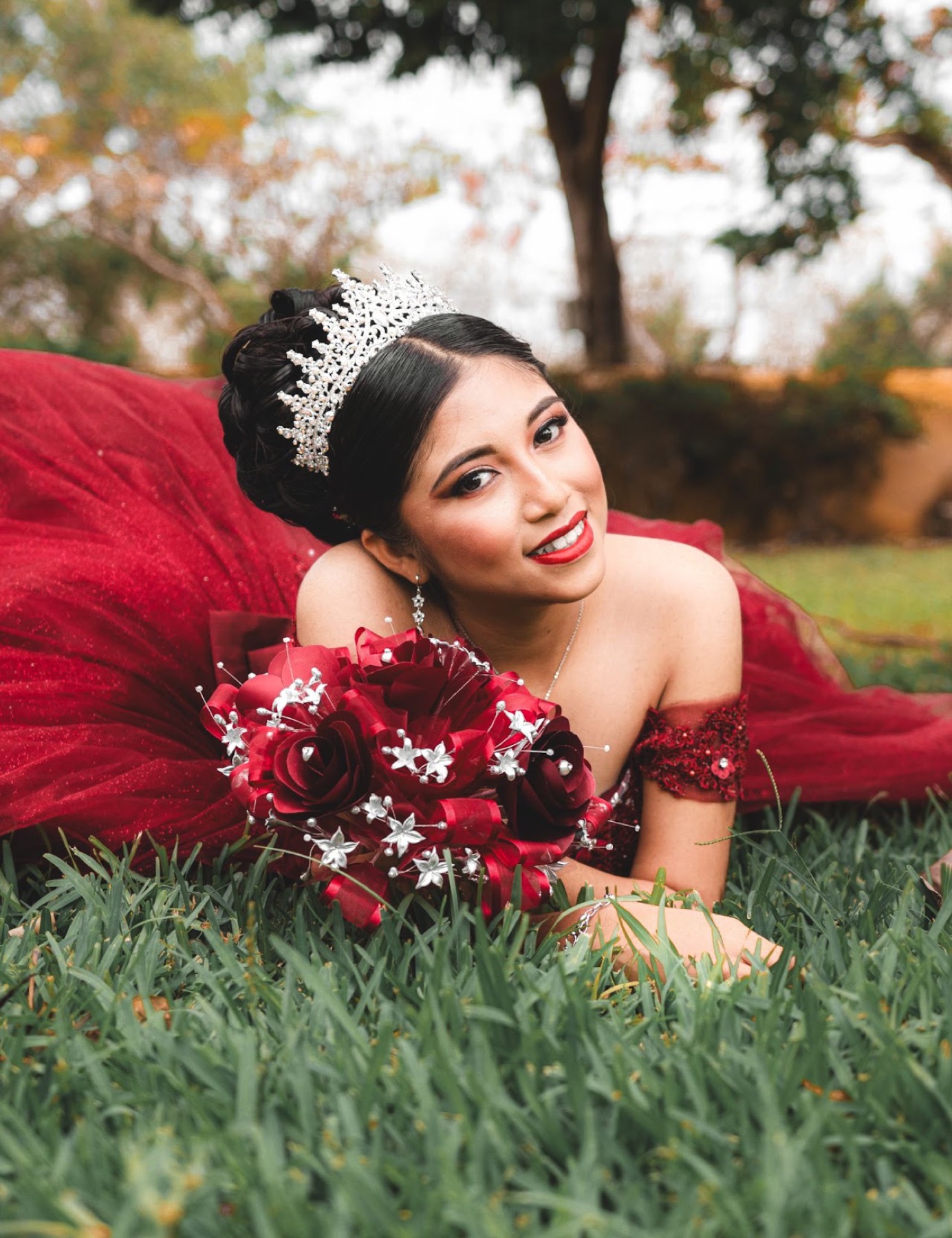 Quinceañera con vestido rojo y ramo recostada sobre el césped