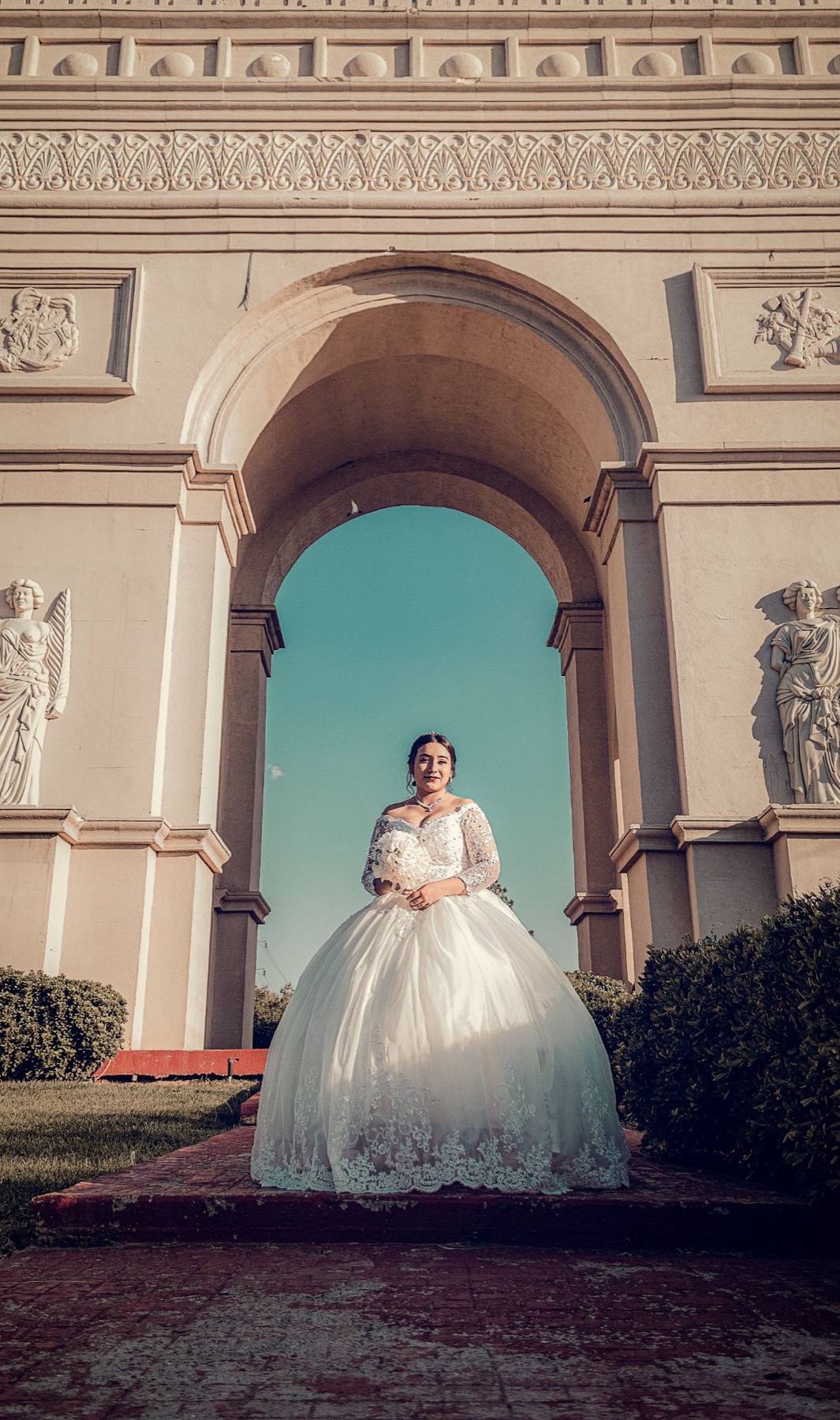 Quinceañera con vestido blanco centrada bajo un arco monumental